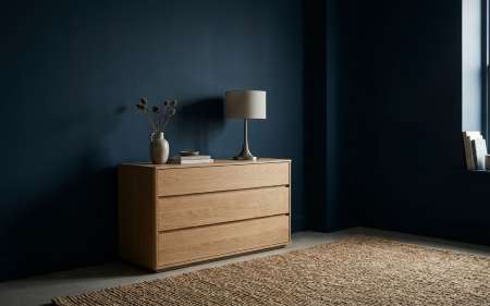 Deep dark blue room interior with modern light oak dresser, table lamp, and textured jute rug